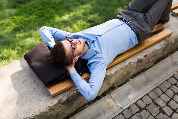 Young handsome man in blue shirt and sunglasses with wireless earphones dreamily lying on bench while spending time in city park