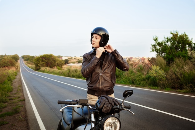 Young handsome man buttoning helmet, standing near his motorbike.