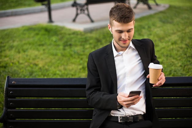 Молодой красивый мужчина в классической черной куртке и белой рубашке с беспроводными наушниками и кофе в руках сидит на скамейке, счастливо пользуясь мобильным телефоном на улице