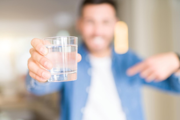 Free photo young handsome man drinking a glass of water at home with surprise face pointing finger to himself