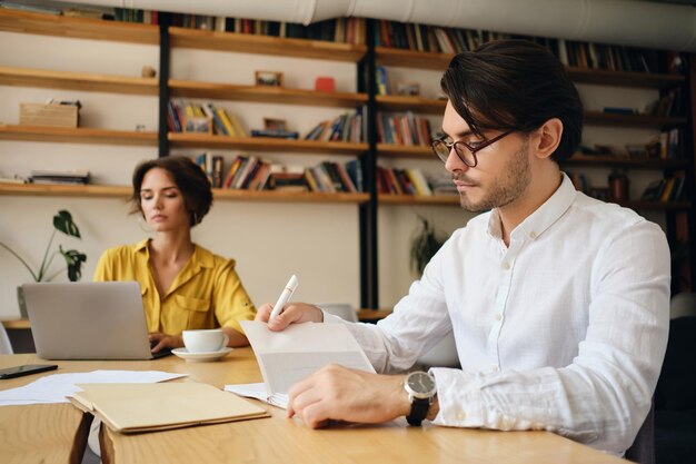 Young handsome man in eyeglasses sitting at the table thoughtfully planning work with colleague on background in modern office