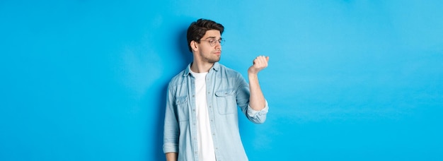 Free photo young handsome man in glasses looking at his fingernails checking manicure standing over blue backgr