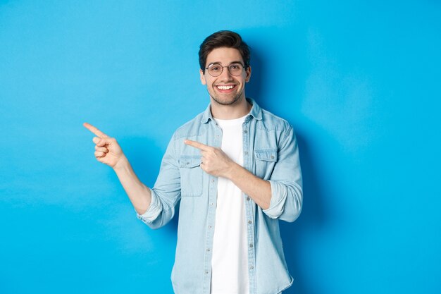안경을 쓴 젊고 잘생긴 남자는 광고를 게재하고 웃고 손가락을 왼쪽으로 가리키며 발표를 하고 파란색 배경에 서 있습니다.