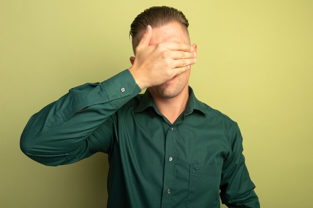 Giovane uomo bello in camicia verde che copre gli occhi con la mano stanca e annoiata in piedi sopra la parete chiara