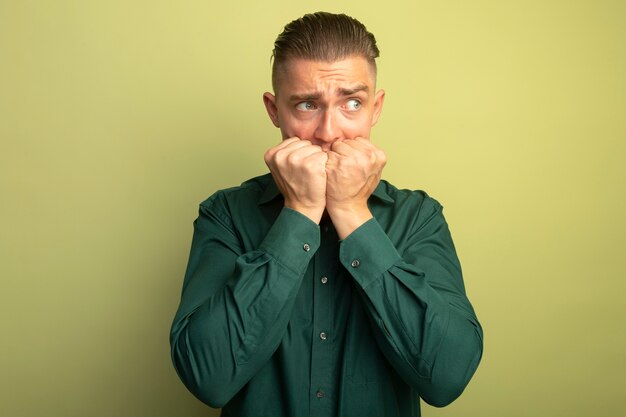 Young handsome man in green shirt looking aside stressed and nervous biting his nails standing over light wall