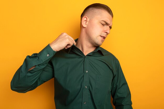 オレンジ色の壁の上に立っている彼の襟を修正する混乱と不満に見える緑のシャツを着た若いハンサムな男