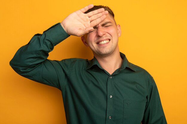 Young handsome man in green shirt looking confused with hand over his head fir mistake forgot important thing standing over orange wall