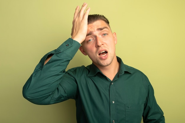 Young handsome man in green shirt looking at front confused with hand on his head forgot important thing standing over light wall