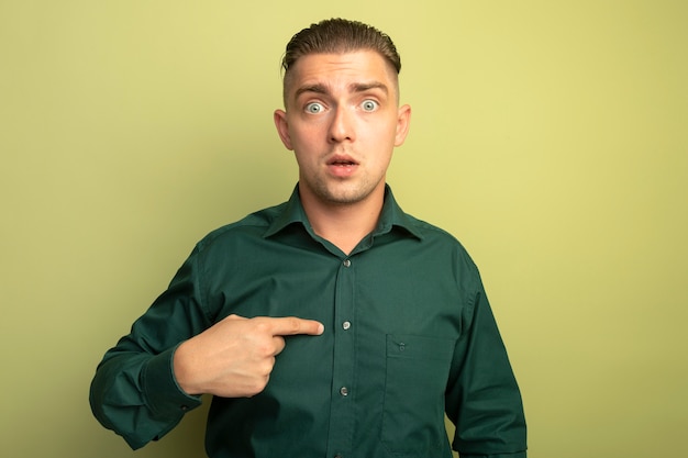 Young handsome man in green shirt pointing at himself being confused standing over light wall