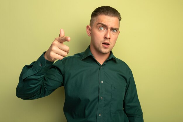 Young handsome man in green shirt pointing with index finger at camera being confused standing over light wall