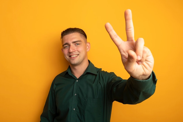 Young handsome man in green shirt smiling cheerfully showing v-sign happy and positive 
