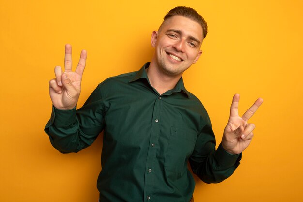 Young handsome man in green shirt smiling cheerfully showing v-sign with both hands 