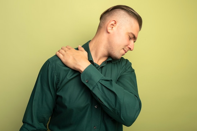 Young handsome man in green shirt touching his shoulder feeling pain standing over light wall
