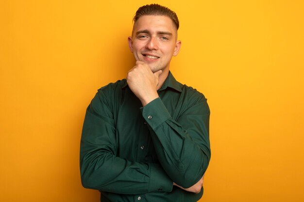 Young handsome man in green shirt with hand on chin smiling confident 