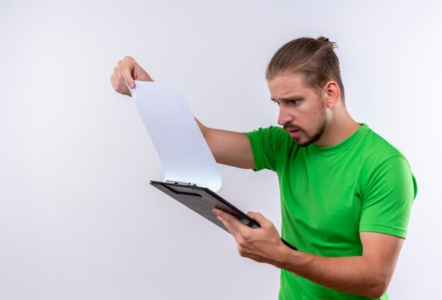 Young handsome man in green t-shirt holding clipboard looking at paper blank surprised and confused standing over white background