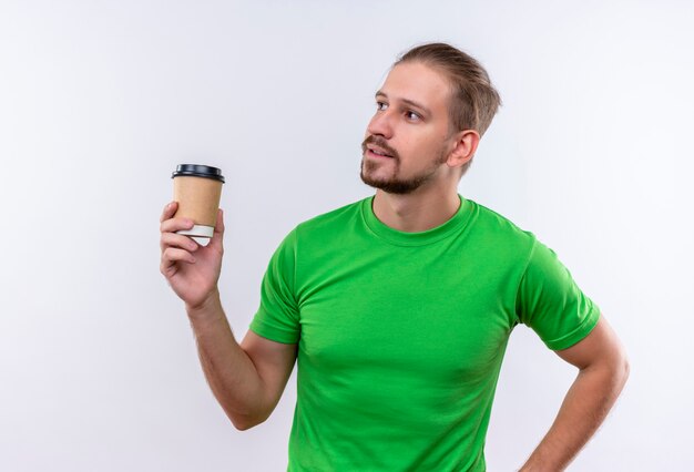 Young handsome man in green t-shirt holding coffee cup looking aside with confident expression on face standing over white background