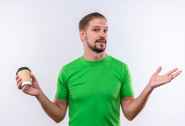 Young handsome man in green t-shirt holding coffee cup looking at camera spreading hands smiling standing over white background