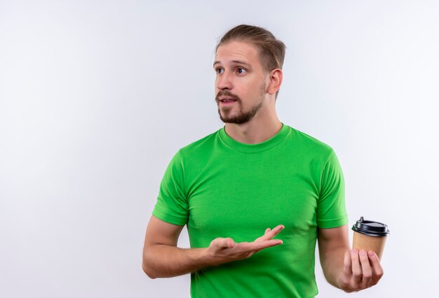 Young handsome man in green t-shirt holding coffee cup looking confused standing over white background