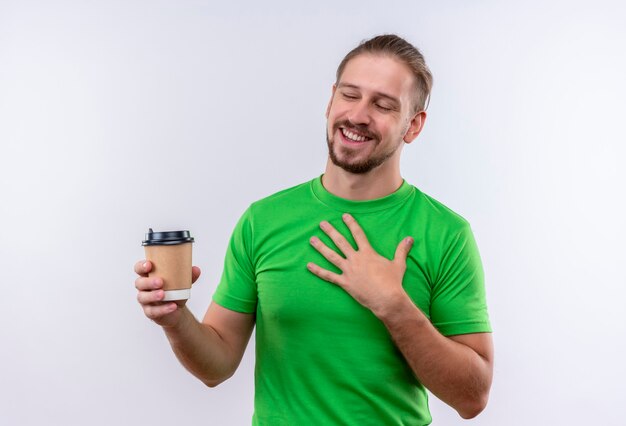 Young handsome man in green t-shirt holding coffee cup smiling cheerfully standing over white background