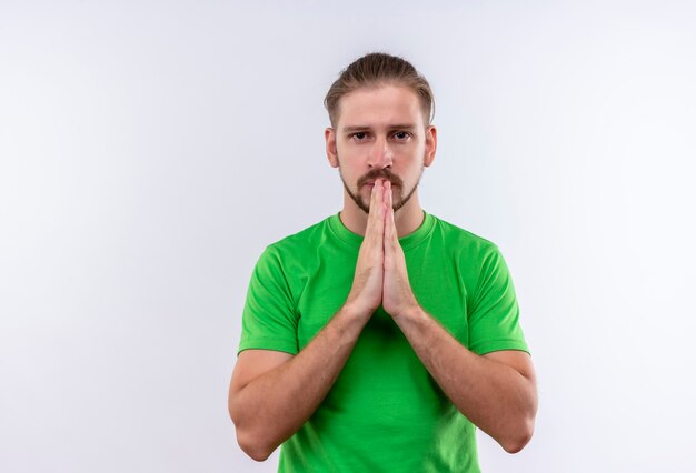 Young handsome man in green t-shirt holding hands together praying with open eyes looking at camera with serious face standing over white background