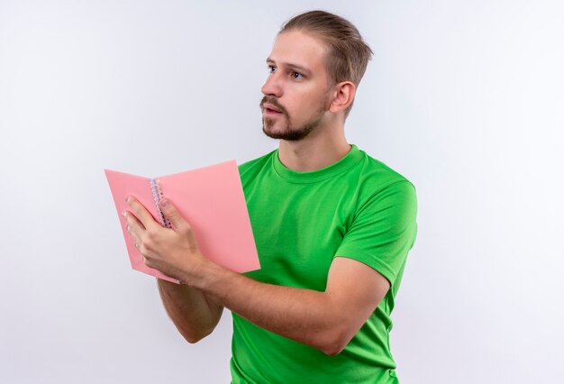 白い背景の上に立っている顔に物思いに沈んだ表情でよそ見ノートを保持している緑のtシャツの若いハンサムな男