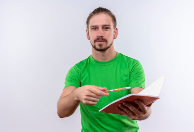 Young handsome man in green t-shirt holding notebook and pencil looking at camera with confident expression standing over white background