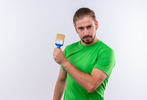 Young handsome man in green t-shirt holding paint brush looking confident standing over white background