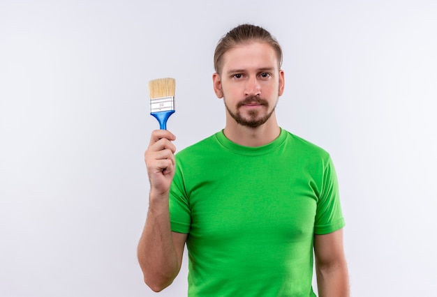 Young handsome man in green t-shirt holding paint brush looking confident standing over white background