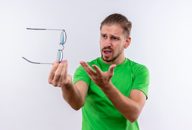 Young handsome man in green t-shirt holding sunglasses in hand looking aside displeased and arguing standing over white background