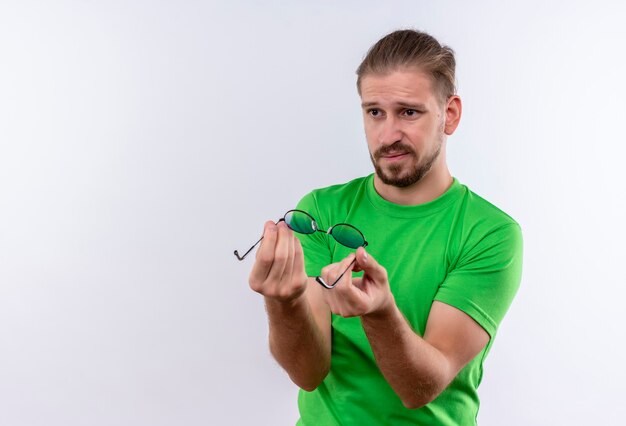 Young handsome man in green t-shirt holding sunglasses in hands looking aside with skeptic expression on face standing over white background