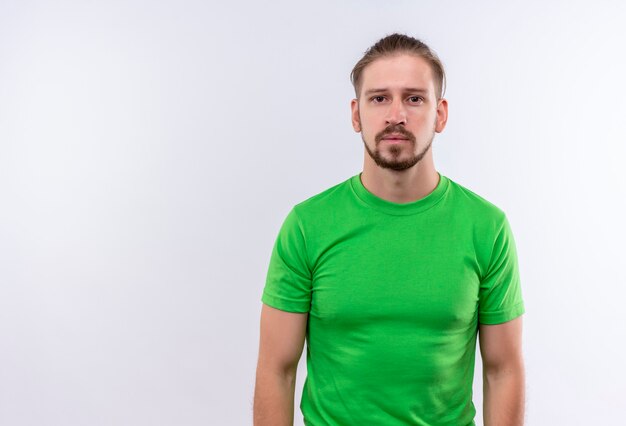 白い背景の上に立っている顔に深刻な自信を持って表情でカメラに緑のtシャツlokingで若いハンサムな男