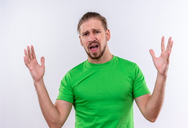 Young handsome man in green t-shirt looking at camera disappointed with raised arms standing over white background