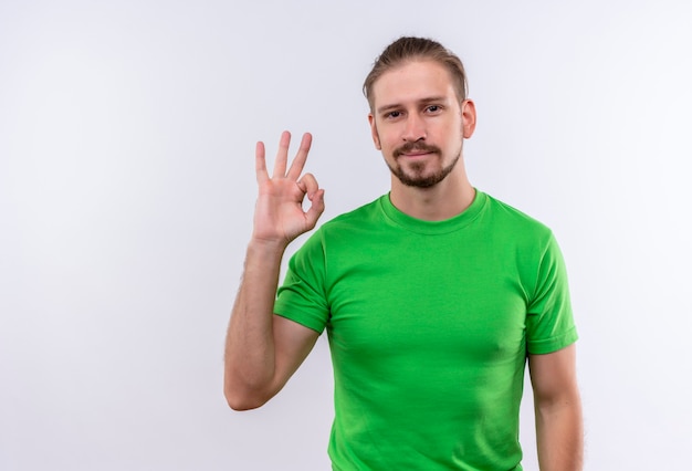 Young handsome man in green t-shirt looking at camera smiling sheerfully doing ok sign standing over white background