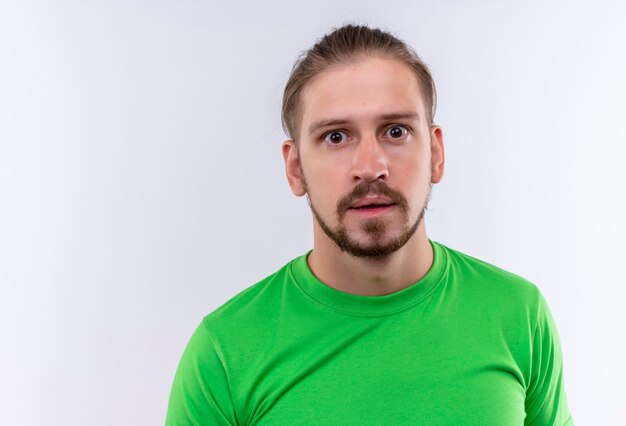 Young handsome man in green t-shirt looking at camera surprised and amazed standing over white background