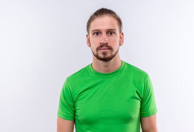 Young handsome man in green t-shirt looking at camera surprised and puzzled standing over white background