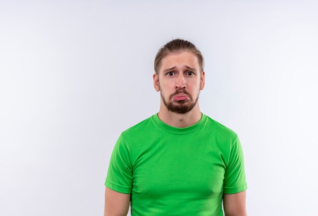 Young handsome man in green t-shirt looking at camera with sad expression on face standing over white background