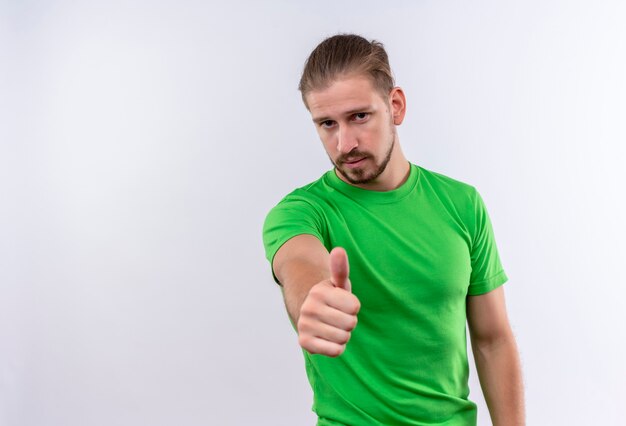 白い背景の上に立ってカメラの親指に自信を持って表示を探している緑のtシャツの若いハンサムな男