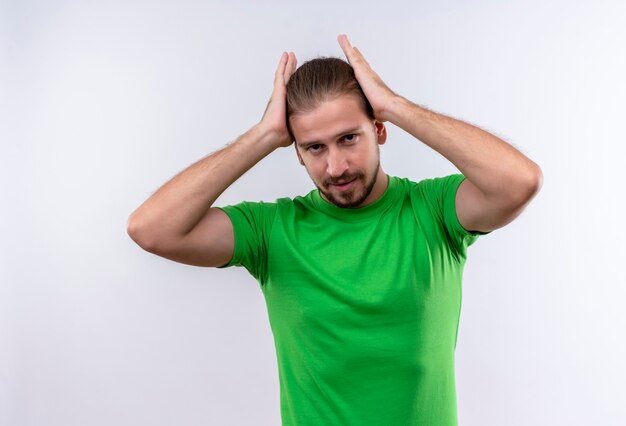 Young handsome man in green t-shirt looking confident touching his perfect hair standing over white background