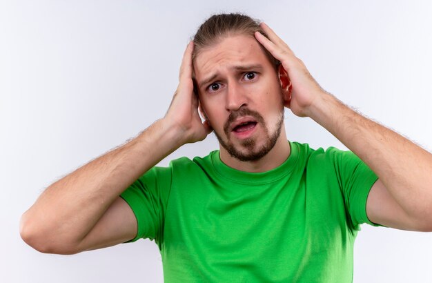 Young handsome man in green t-shirt looking surprised and confused touching his head standing over white background