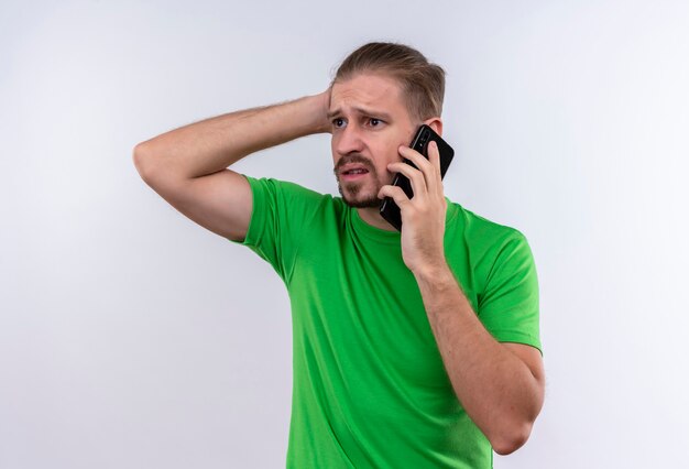 Young handsome man in green t-shirt talking on mobile phone looking confused and very anxious standing over white background