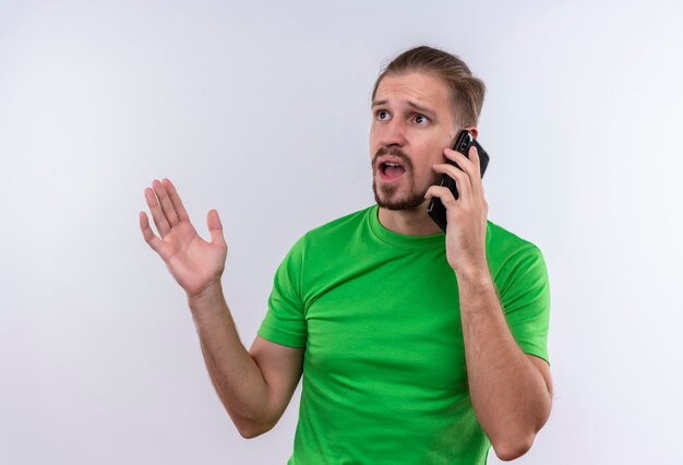 Young handsome man in green t-shirt talking on mobile phone looking confused and very anxious standing over white background