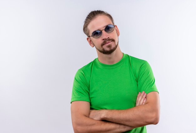 Young handsome man in green t-shirt wearing glasses looking confident standing over white background