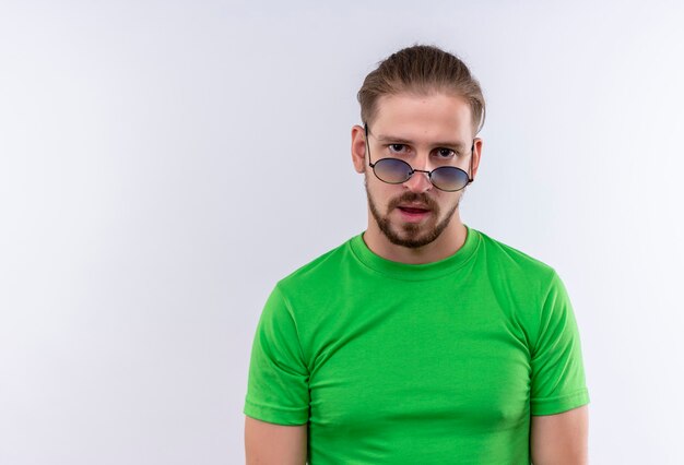 Young handsome man in green t-shirt wearing sunglasses looking confident self-satisfied standing over white background