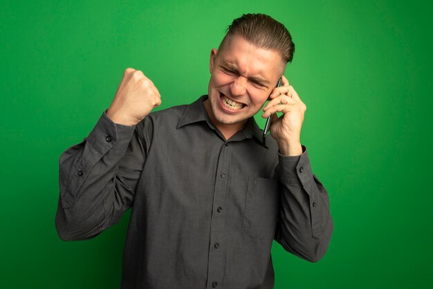 Foto gratuita giovane uomo bello in camicia grigia che stringe il pugno felice ed eccitato mentre parla al telefono cellulare in piedi sopra la parete verde