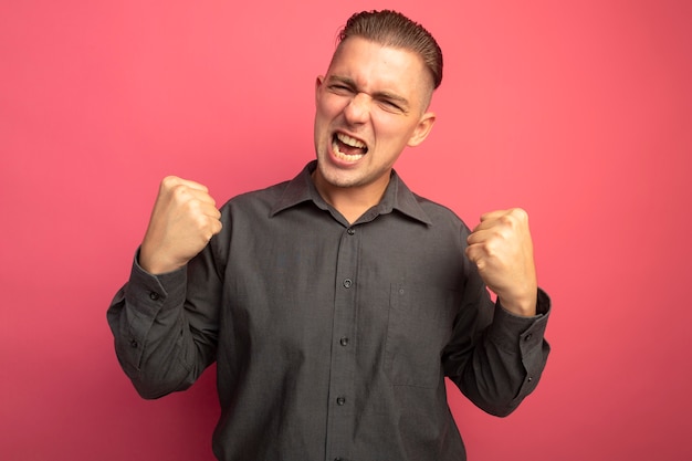 Free photo young handsome man in grey shirt clenching fists happy and excited shouting standing over pink wall