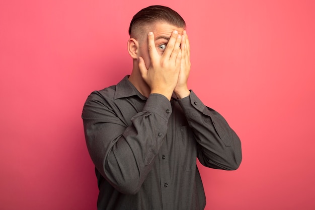 Free photo young handsome man in grey shirt covering eyes with hands looking through his fingers standing over pink wall