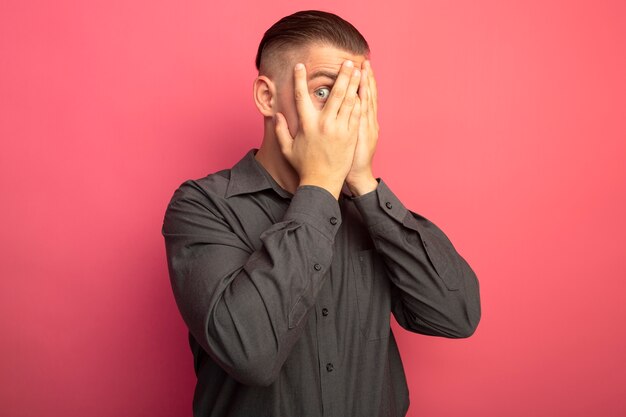 Young handsome man in grey shirt covering eyes with hands looking through his fingers standing over pink wall