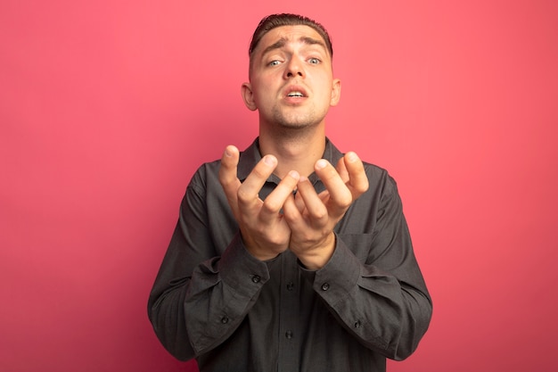 Young handsome man in grey shirt holding arms out like begging with hope expression standing over pink wall