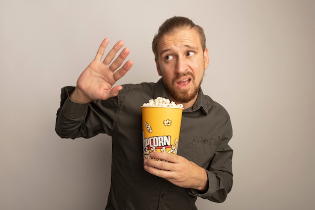 Young handsome man in grey shirt holding bucket with popcorn looking aside worried and confused holding hand out like defense gesture 