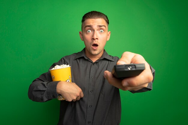 Young handsome man in grey shirt holding bucket with popcorn using tv remote looking at front amazed standing over green wall